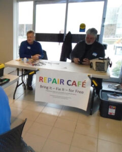 Two men sitting behind a table with a sign stating Repair Cafe. 