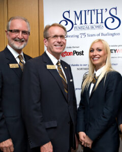 2 team members from Smith's Funeral Home in Burlington, standing in front of a business sign.