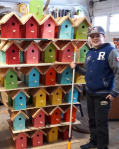 Young boy standing beside a stack of multi coloured birdhouses.